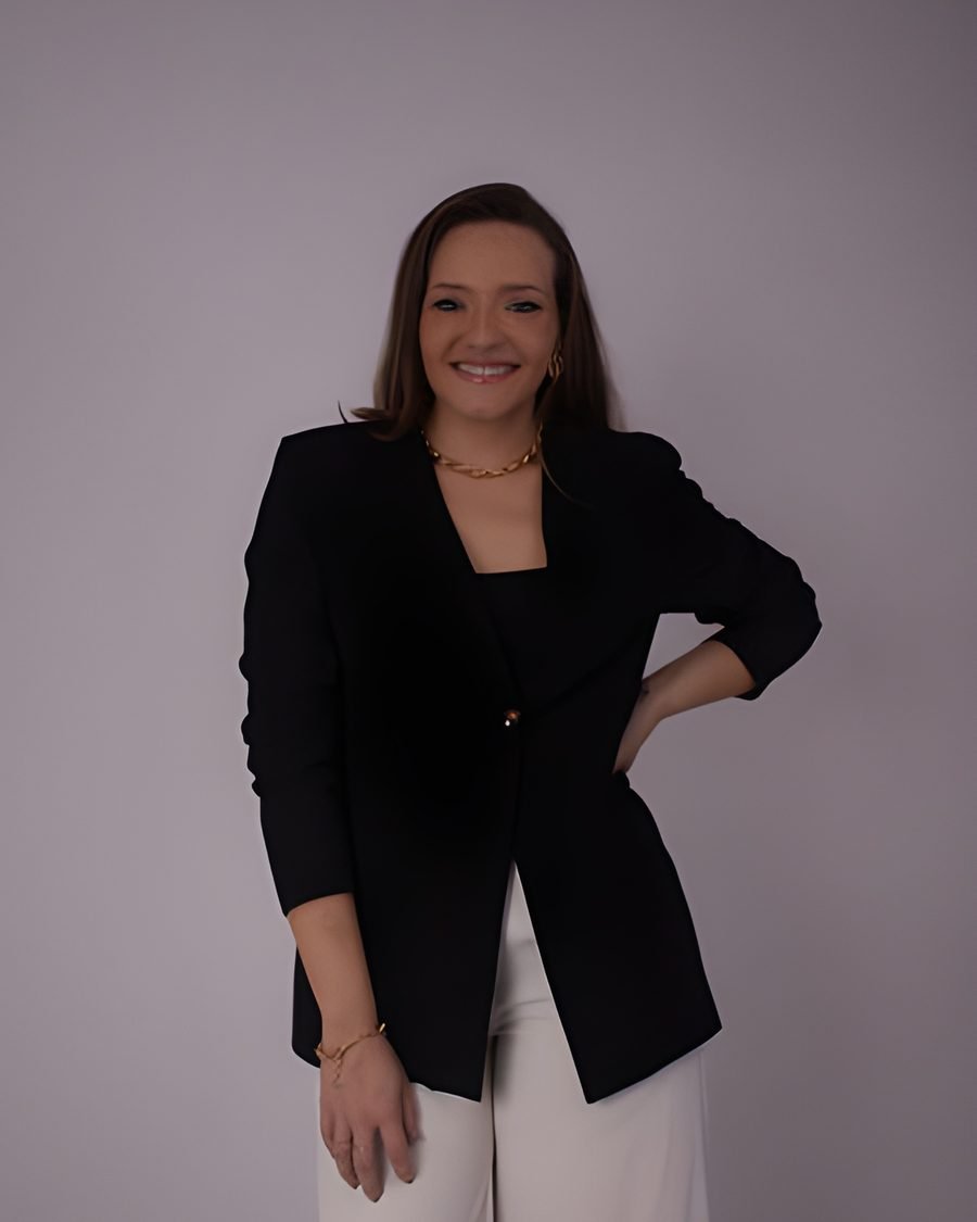 Mayalle Fernandes em estúdio, sorrindo, de blazer preto, colar dourado e calça clara, com a mão na cintura
