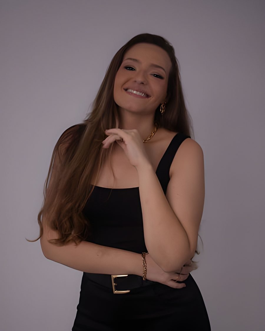 Mayalle Fernandes em estúdio, sorrindo, de regata preta com cinto e acessórios dourados
